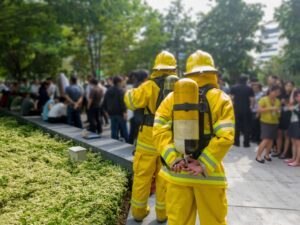 Disaster Response Two firefighters dressed in yellow suits, seen from the back, standing in front of a crowd during an emergency drill or event.