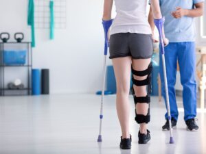 Rehabilitation A person wearing a leg brace and using crutches during a physical therapy session, with a therapist standing nearby.