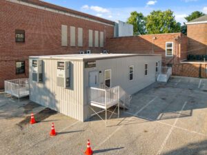 A modular building with a metal exterior, labeled ‘Nadler Modular,’ situated in a parking lot adjacent to a brick building, with orange traffic cones marking the area.