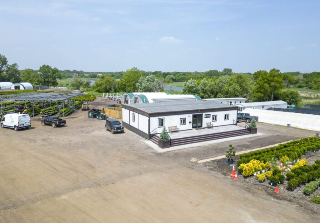 Aerial view of a large nursery or garden center featuring a central building with parking area and various greenhouses. Surrounding the building are rows of neatly arranged plants and trees. A few vehicles are parked nearby under a clear, blue sky.