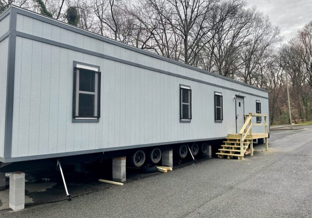 A light gray trailer with black trim is supported by blocks and wheels on a paved surface near leafless trees. The trailer has several windows, and a small wooden staircase leads up to a door. The sky appears overcast.