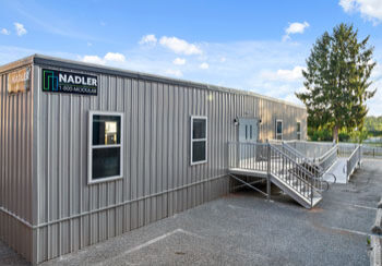 Nadler-Modular-Portable-Classroom-Exterior (1) A modular building with a metal exterior, featuring a sign that reads ‘Nadler Modular,’ with a ramp leading to the entrance.