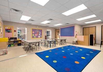 Nadler-Modular-Portable-Classroom-Interior A brightly lit classroom with a blue carpet featuring colorful dots in the center. Surrounding the carpet are small tables and chairs. The walls display various educational posters and a bulletin board. Shelves with toys and supplies are on the left side.